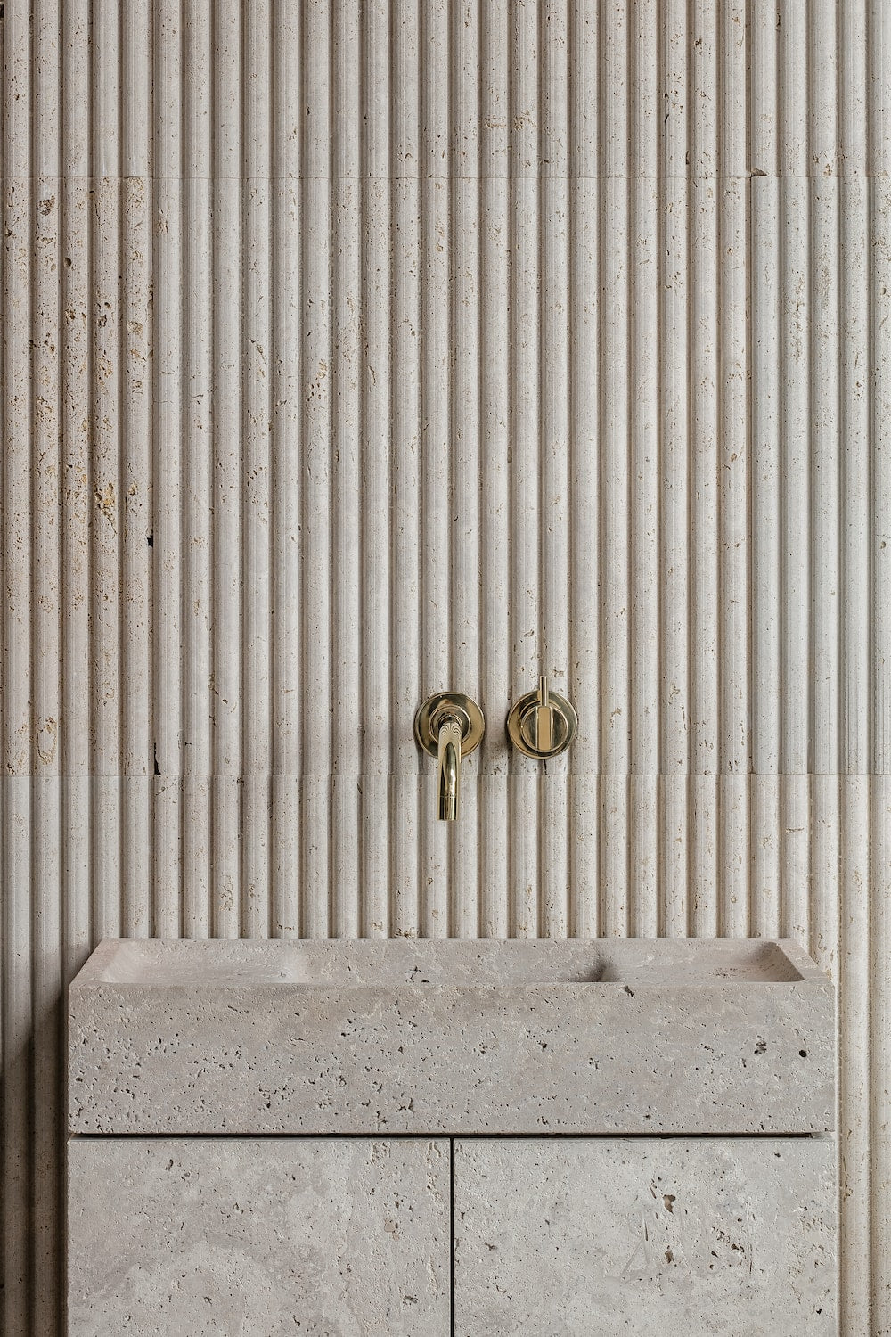 Stone bathroom vanity with integrated sink and brass wall-mounted faucet against fluted stone wall